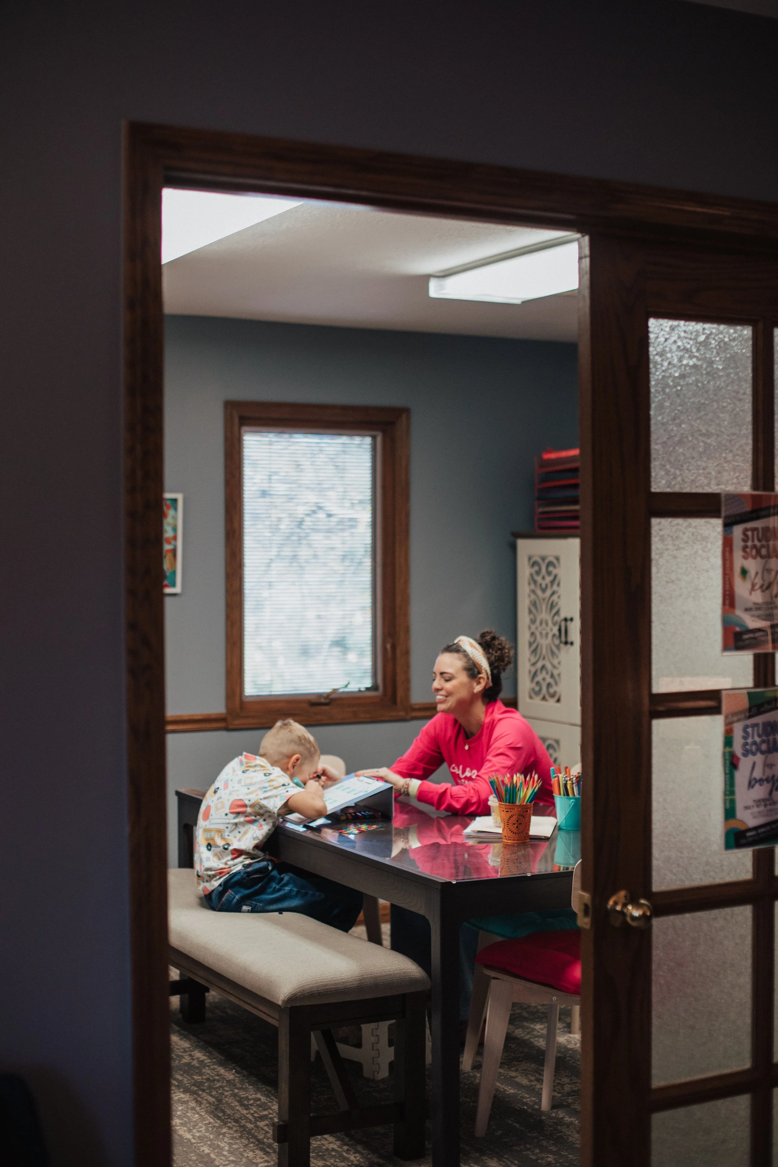 Kids occupational therapist in Columbus, OH and young boy sitting at a table in a room, engaging in a learning activity with art supplies and a book, viewed through a doorway.