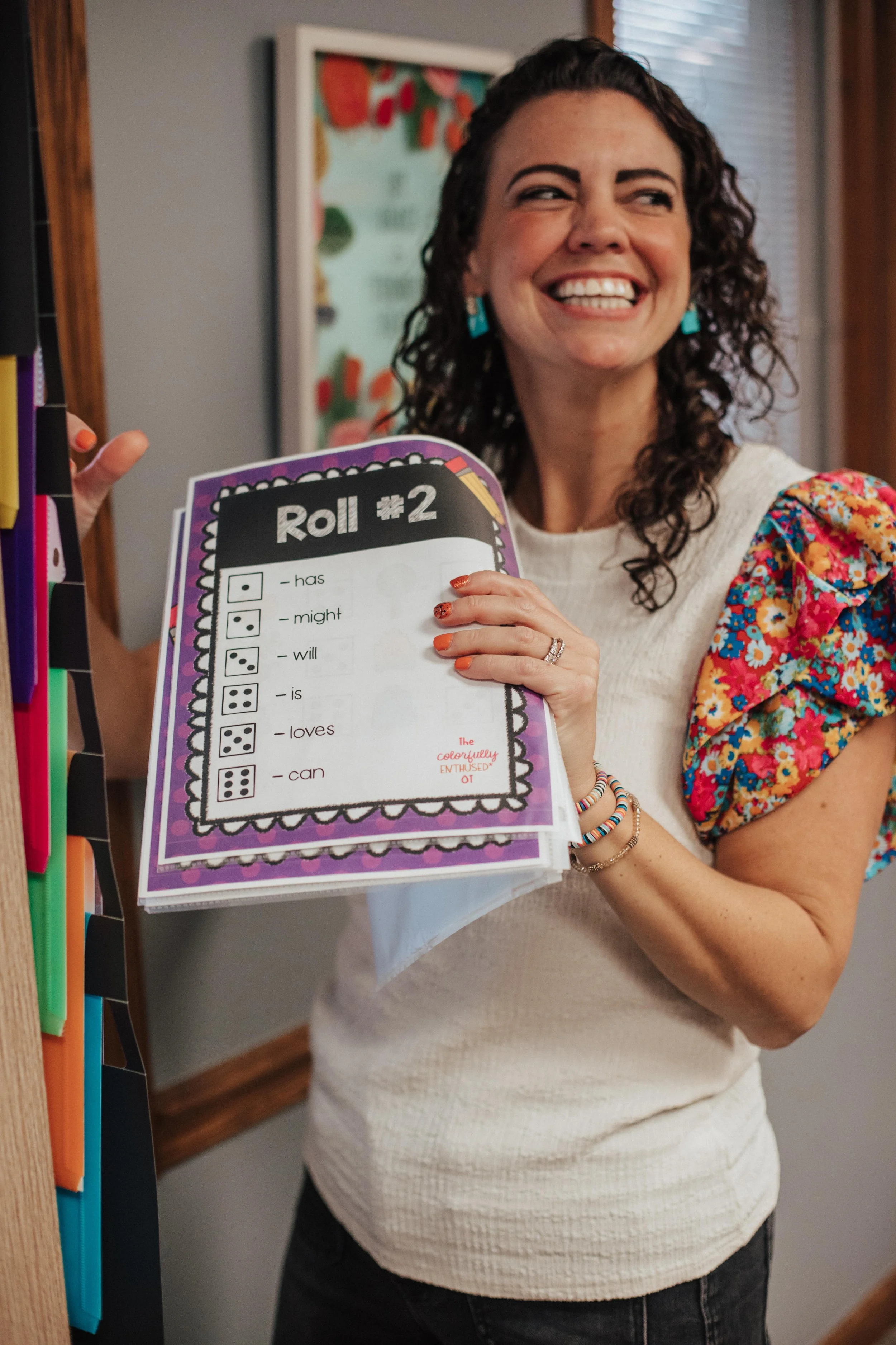 Occupational therapist in Westerville, OH holding a colorful educational chart titled 'Roll #2' with dice and phrases like 'has,' 'might,' 'will,' 'is,' 'loves,' and 'can'.