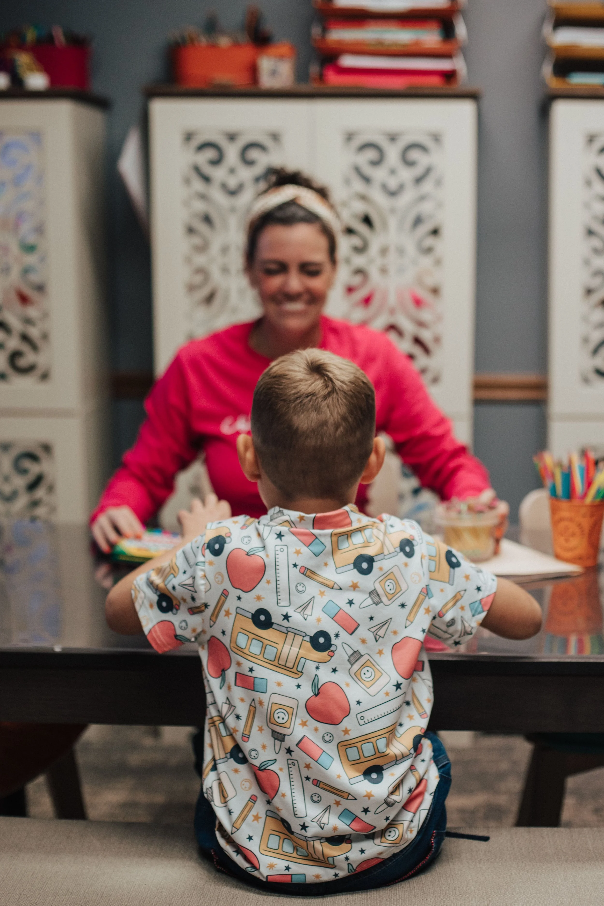 A young boy with short hair wearing a colorful shirt with school bus and apple patterns, sitting at a table across from his occupational therapist in Westerville, OH, in a room with decorative cabinets and shelves with books and art supplies.
