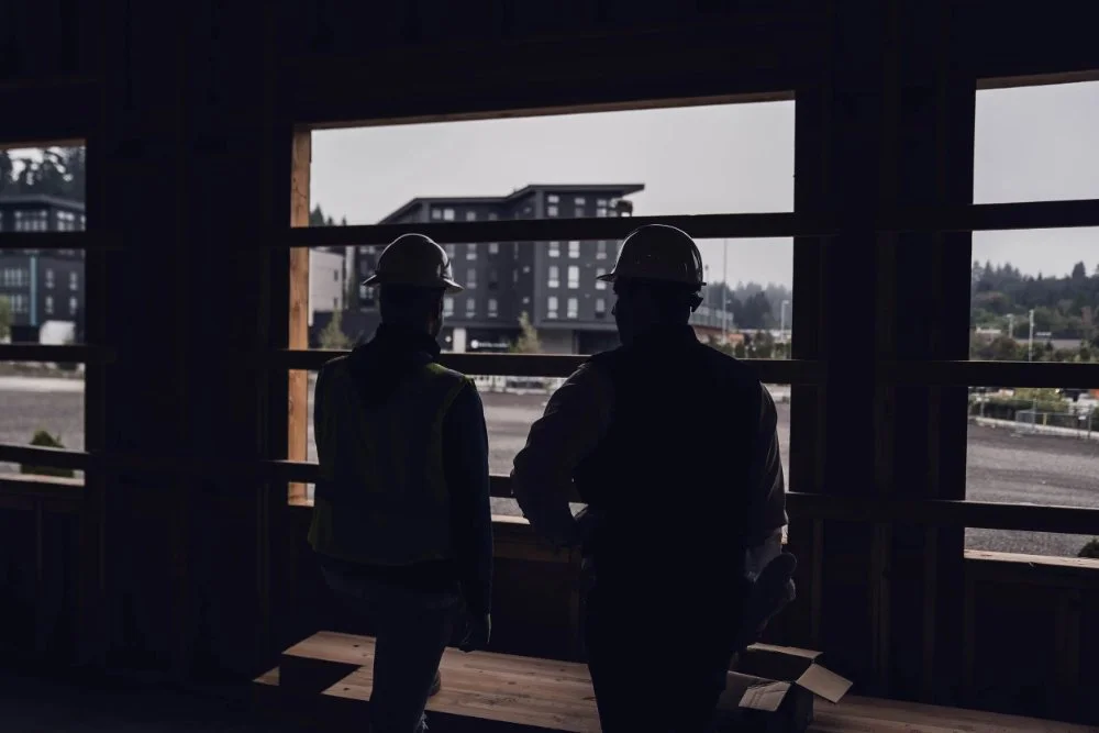 Two construction workers wearing hard hats stand inside a building under construction, observing the exterior through a large rectangular window, with a modern building and construction site visible outside.