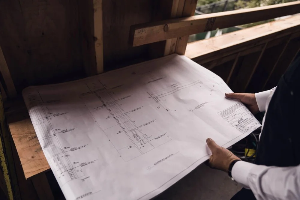 Person in suit holding large construction blueprint on wooden table in building under construction.