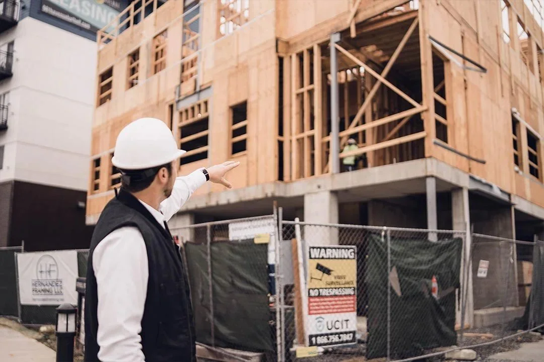 A construction site with a man wearing a hard hat and vest pointing towards a building under construction with wood framing.
