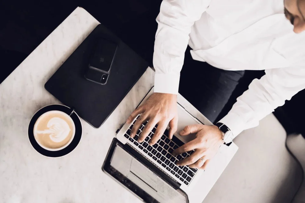 Person in white shirt working on a laptop at a white marble table, with a coffee cup and a smartphone nearby.