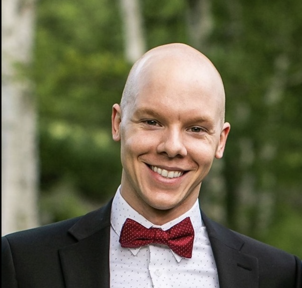 A smiling man with a shaved head, wearing a black tuxedo with a white shirt and red polka dot bow tie, standing outdoors with green trees in the background.