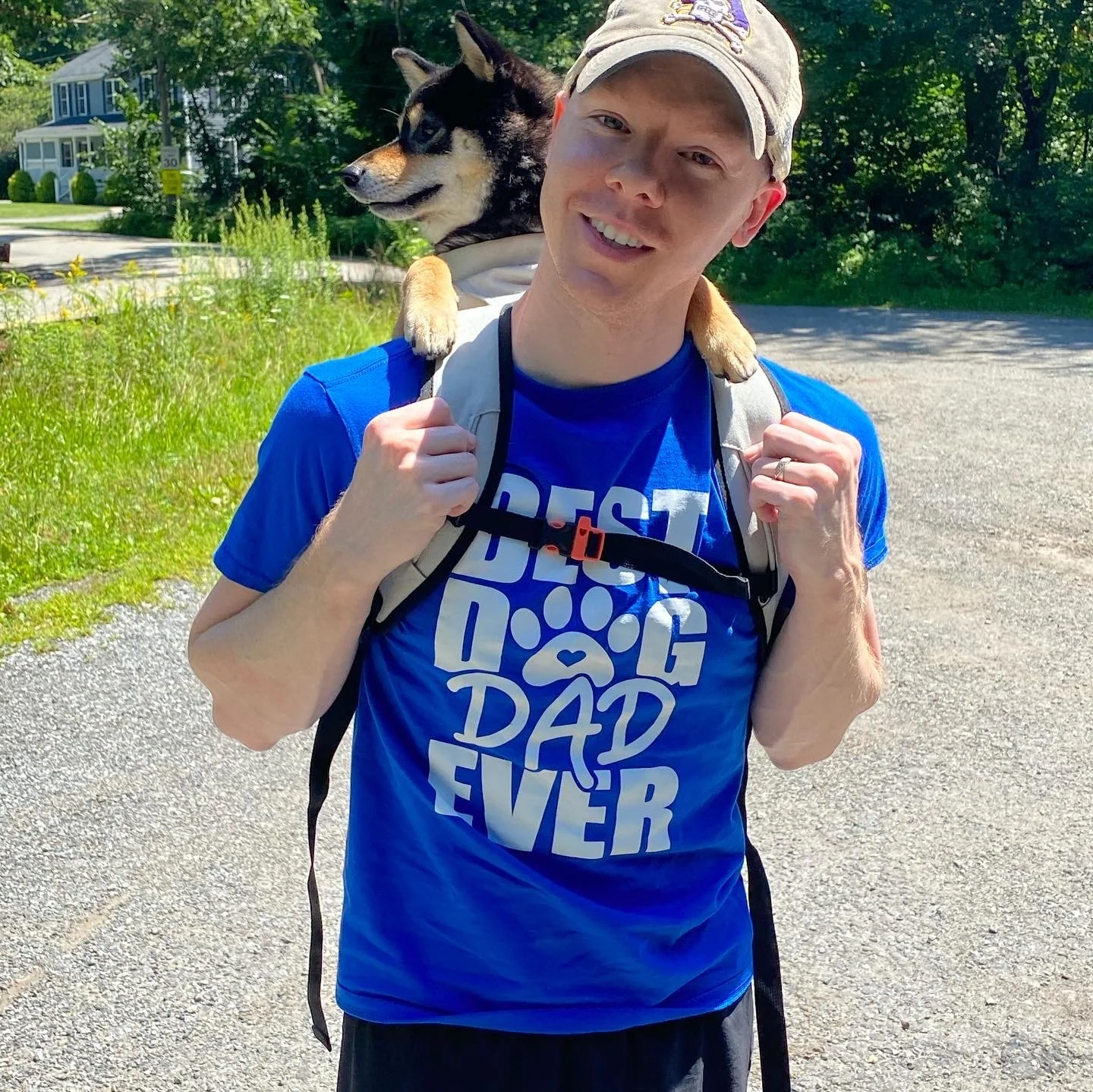 Man wearing a blue T-shirt with the text 'Best Dog Dad Ever' and a paw print, carrying a dog on his shoulders during daytime outdoors.