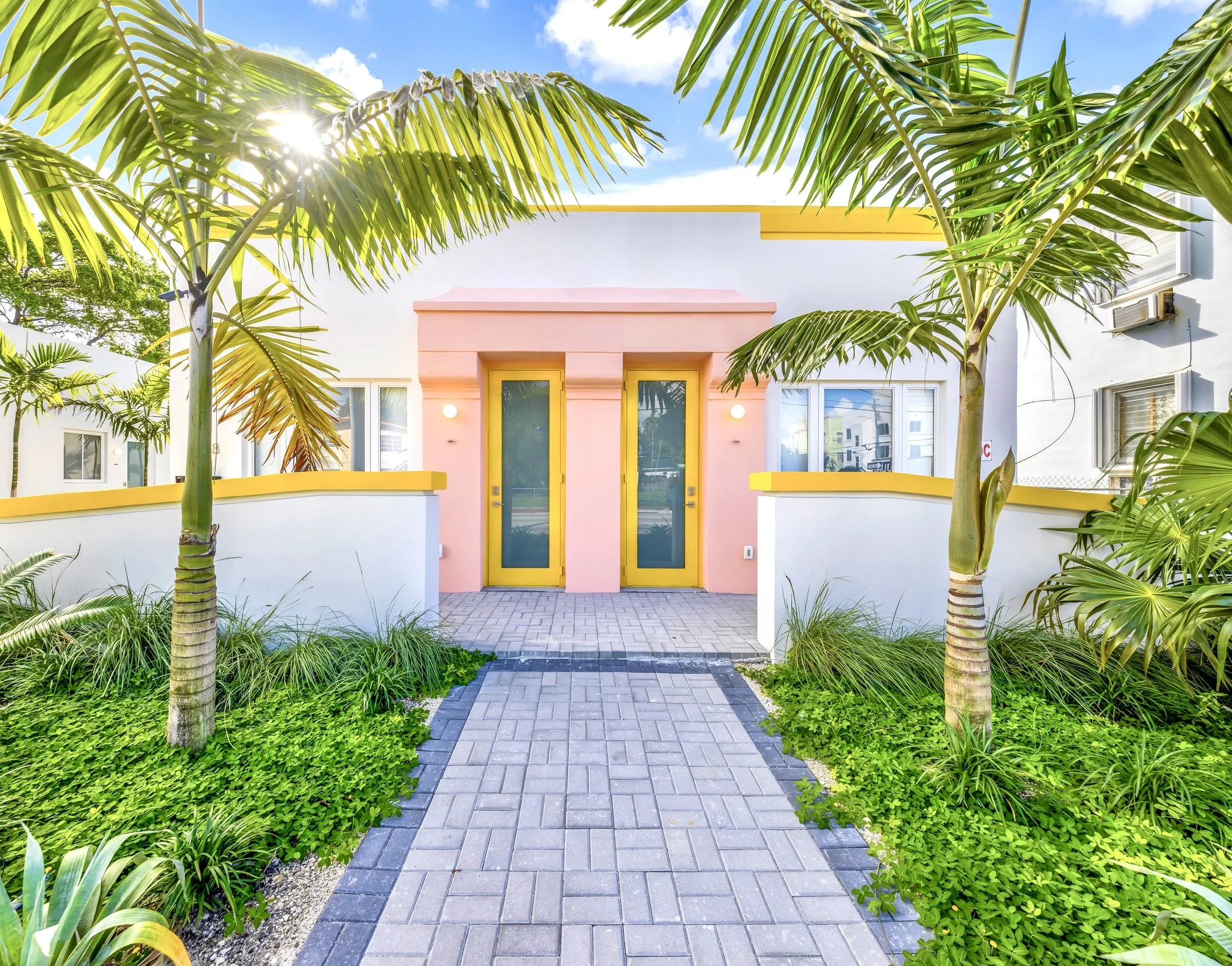 Front view of a modern house with white and pink facades, yellow accents, glass doors, surrounded by palm trees and lush greenery, under a blue sky.