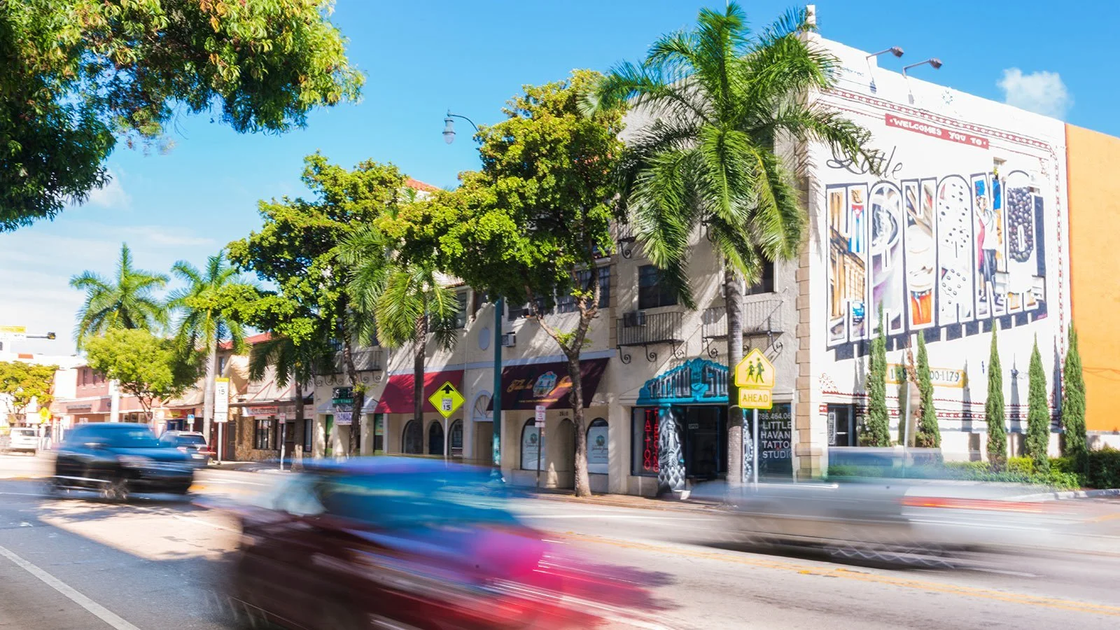 Street scene with blurred cars driving past storefronts and colorful building murals, lined with trees