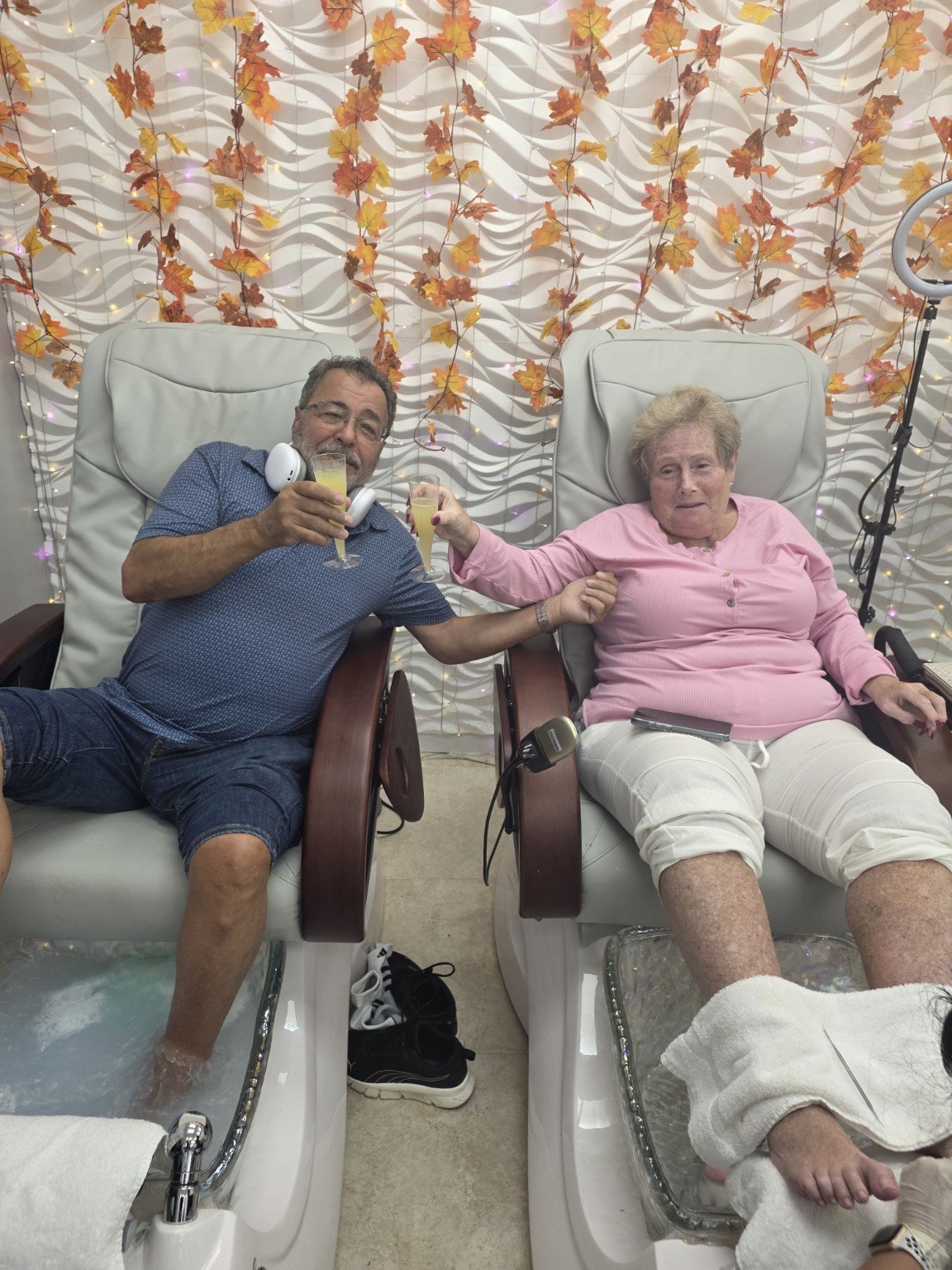 An elderly woman and a middle-aged man are sitting in separate pedicure chairs, holding hands and toasting with champagne glasses. The background has a decorative wall with autumn leaves and string lights, creating a festive atmosphere.