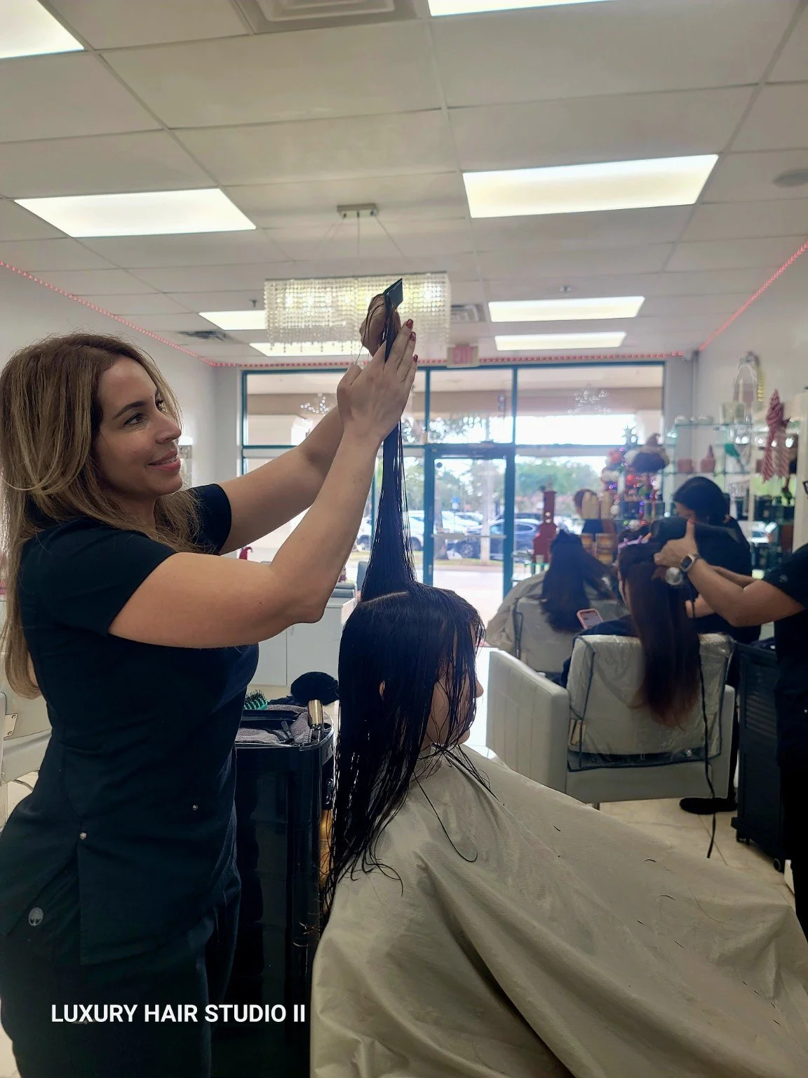 A hairstylist with long, light brown hair in a black shirt is lifting a section of a client's wet, dark brown hair in a hair salon. The client is seated in a chair covered with a beige cape. There are other people and hairstyles visible in the backgr