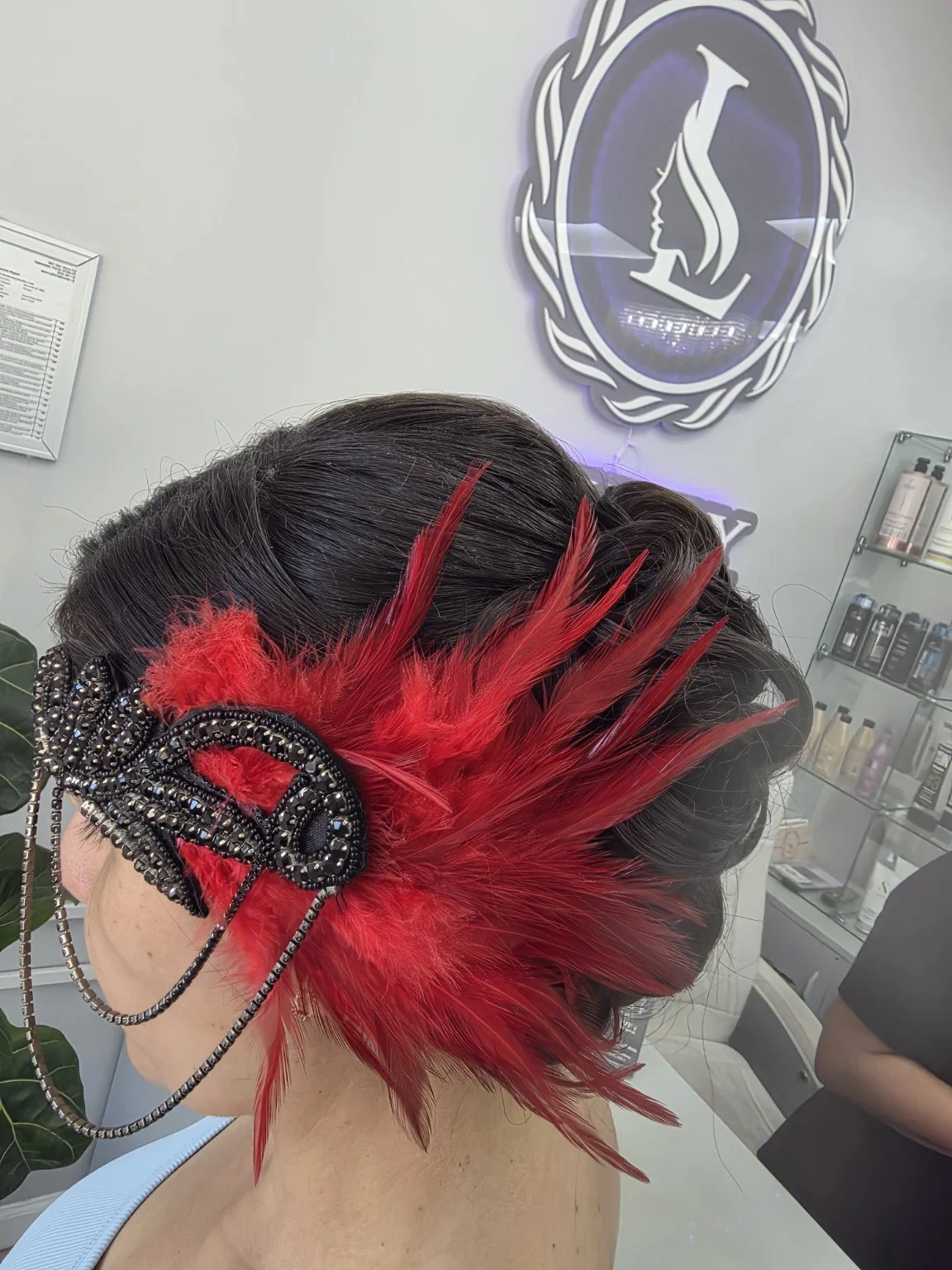 Woman with styled black hair accessorized with a large, black bejeweled hair clip and vibrant red feathers, inside a salon with hair products on shelves and a logo on the wall.