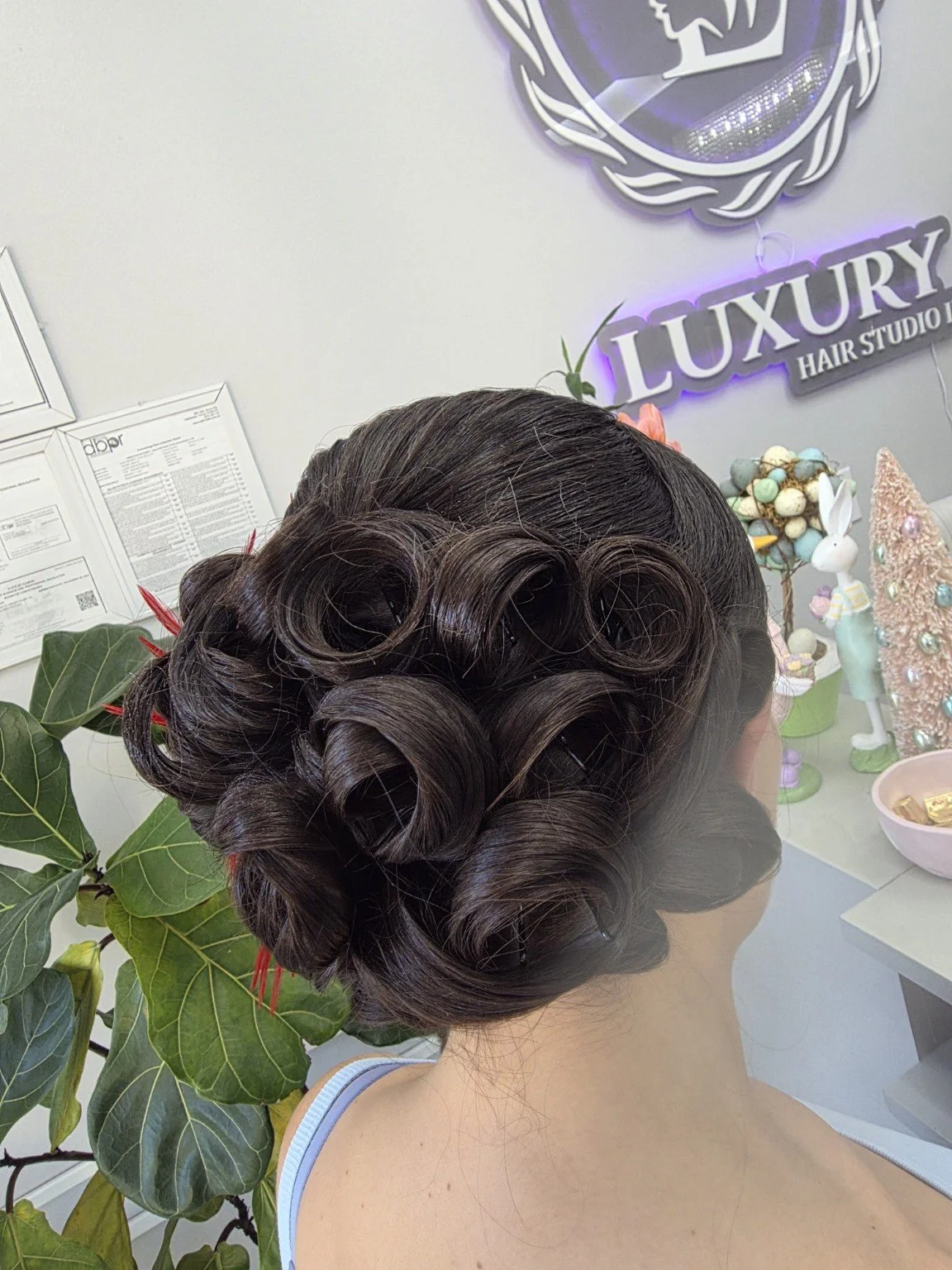 A woman with dark brown hair styled in vintage finger waves and pin curls, sitting in a hair salon with Easter decorations in the background.