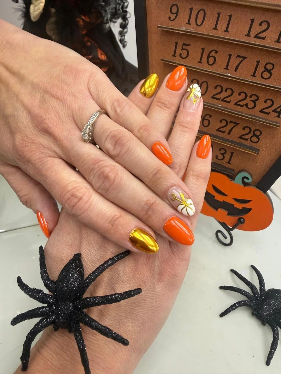 Two hands with decorated nails, one wearing a diamond ring, in front of a Halloween-themed décor including a wooden calendar with October dates, a carved pumpkin with a face, and glittery plastic spiders.