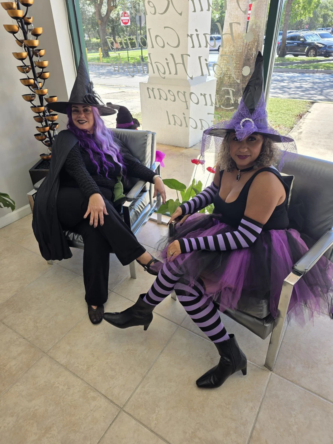 Two women dressed as witches sitting on chairs inside a building near a large window with a view of parked cars and trees outside. One woman has purple hair and a black witch hat, the other has curly hair and a purple and black witch hat. There are p