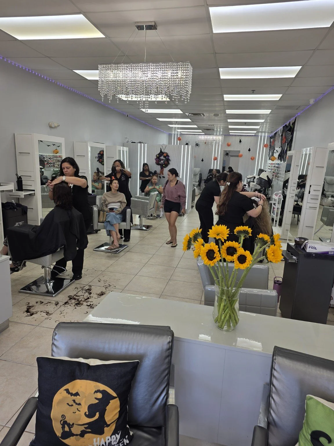 Interior of a salon with people getting haircuts and treatments, decorated with Halloween themed ornaments, including sunflowers in a vase on the foreground table and a black pillow with a Halloween design on a chair.