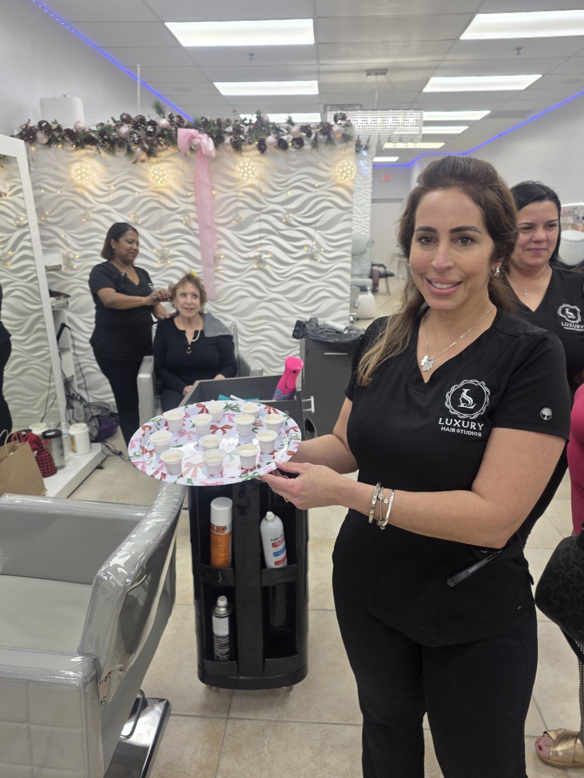 A woman wearing a black shirt with a logo that says 'Luxury Hair Studio' is holding a tray with small cups of what appears to be a beverage, in a salon decorated for Christmas with holiday lights and decorations.