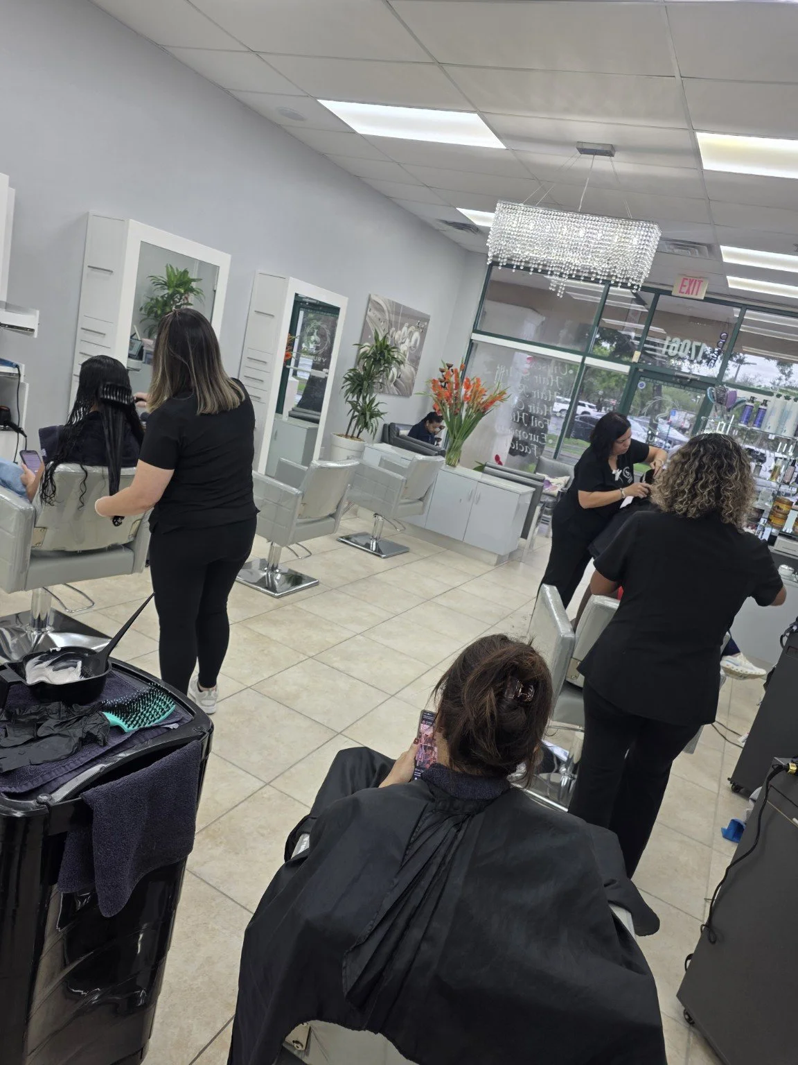 Interior of a hair salon with several clients getting hair treatments, stylists working, and a woman seated at a station using a smartphone.