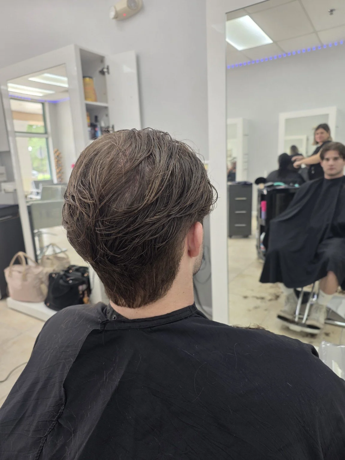 Back of a man sitting in a hair salon chair, getting a haircut, with a mirror reflecting the stylist and another customer in the background.