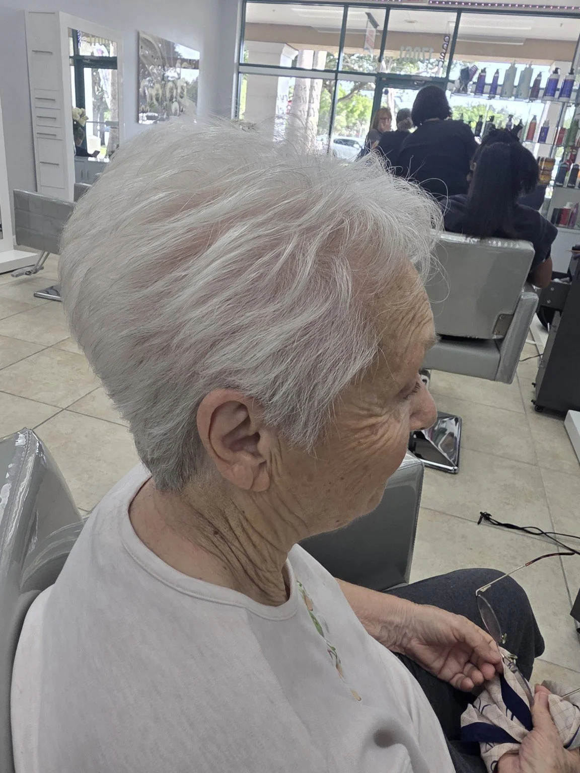Older woman with white hair sitting in a salon, seen from the side, with styling chairs and other clients in the background.
