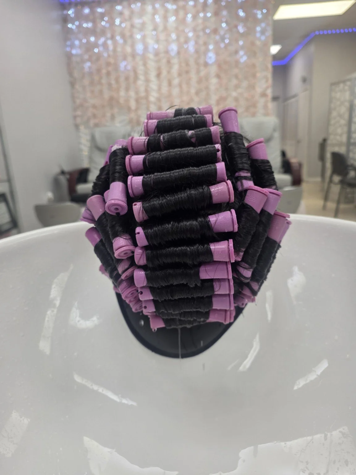 Multiple black hair rollers with pink ends arranged on a hair mannequin head, set in a salon or beauty salon with blurred background.