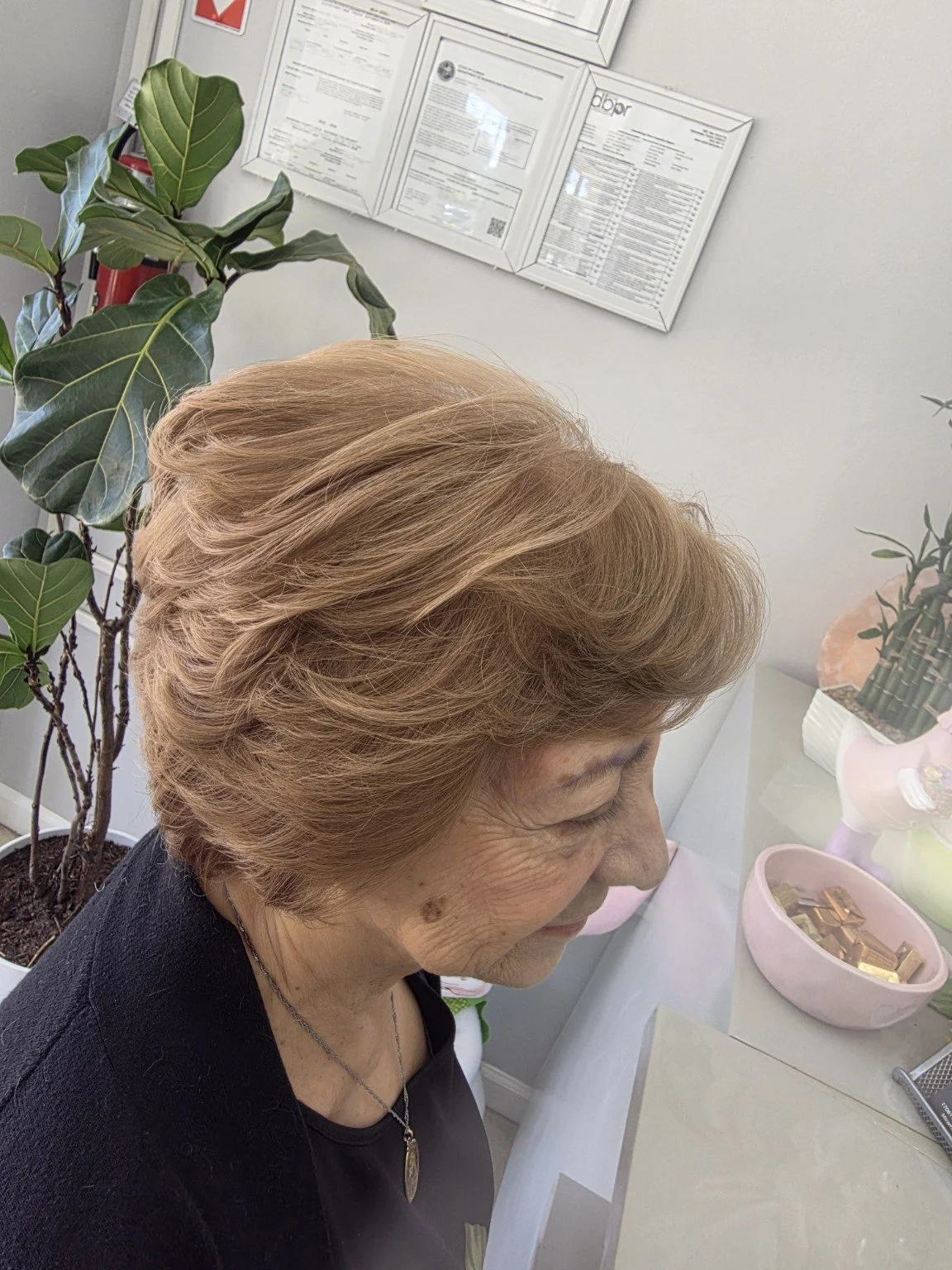 A woman with styled, light brown, wavy hair is smiling and looking down. She is wearing a black top and a necklace. There are plants and framed documents on the wall behind her, and a table with a pink bowl filled with chocolates or candies, and some