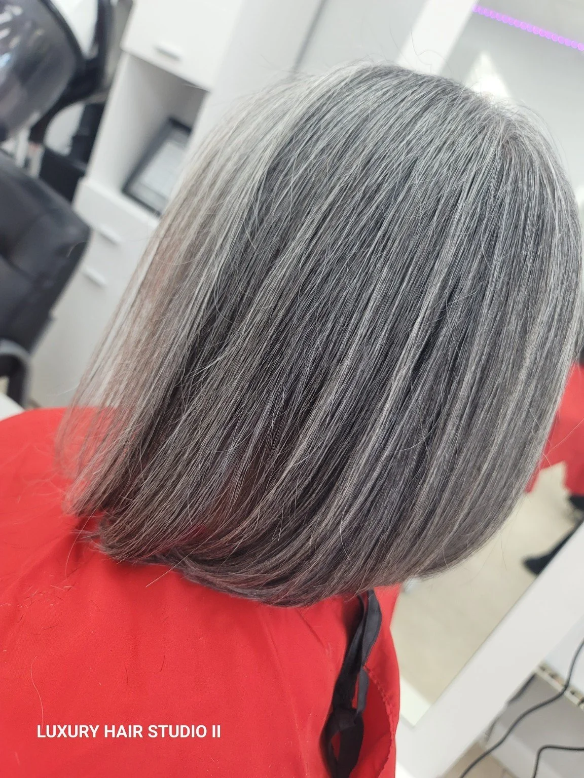 Side view of a woman's head with straight, shoulder-length gray hair, seated in a salon with white cabinets and salon equipment in the background.