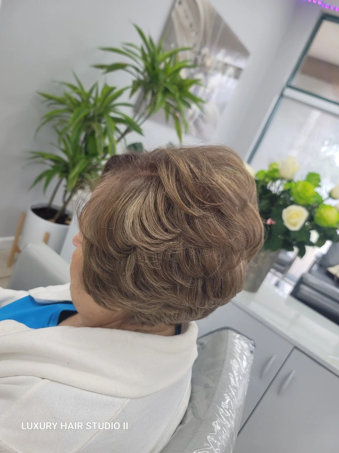 Side view of a woman with short, styled, light brown hair, sitting in a salon chair. Behind her are large green plants and a display of white and yellow flowers in the background.