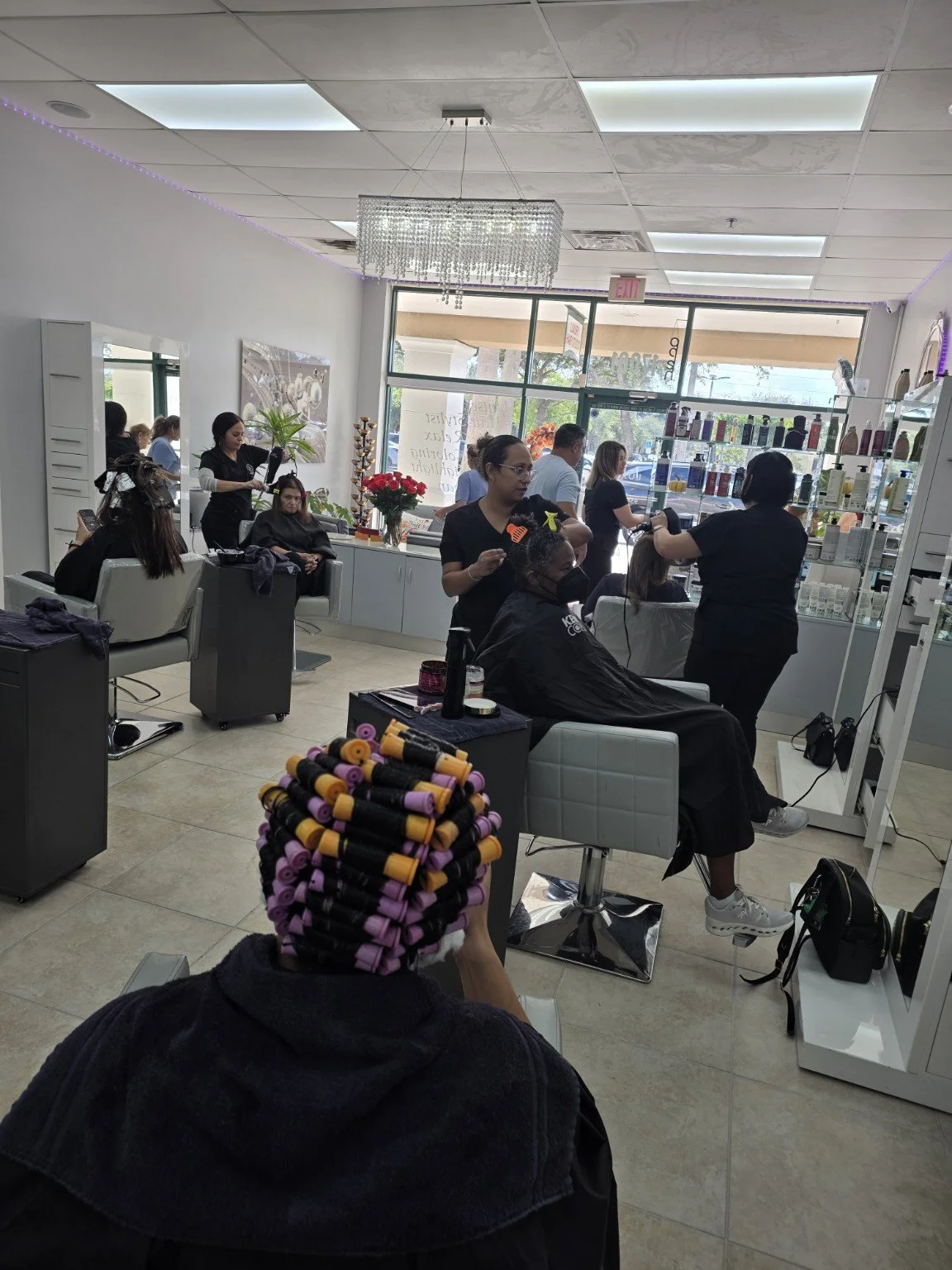 Interior of a hair salon with multiple clients and stylists. Several clients are getting hair treatments; one has colorful curlers in her hair. Hair products are displayed on shelves at the front. The salon has large windows letting in natural light.