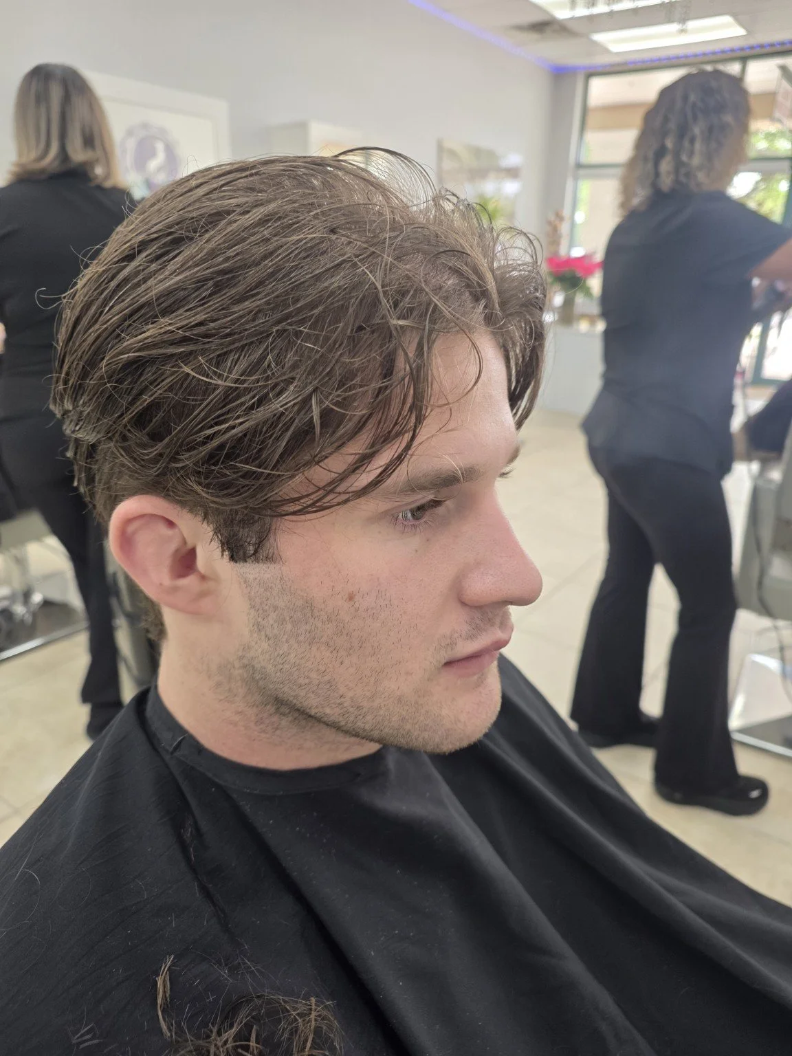 Young man with professional hairstyle sitting in a salon chair, wearing a black cape, while two hairstylists working in the background.