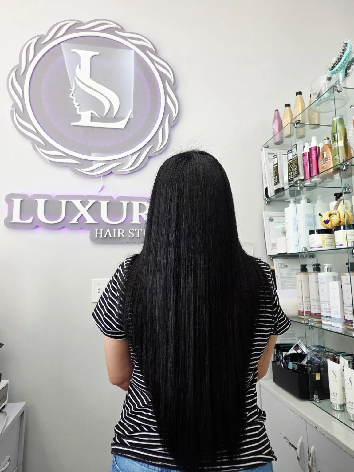 Back of a woman with long, straight black hair standing in a hair salon. The salon has a sign that reads 'Luxury Hair Studio' on the wall and shelves with hair products.