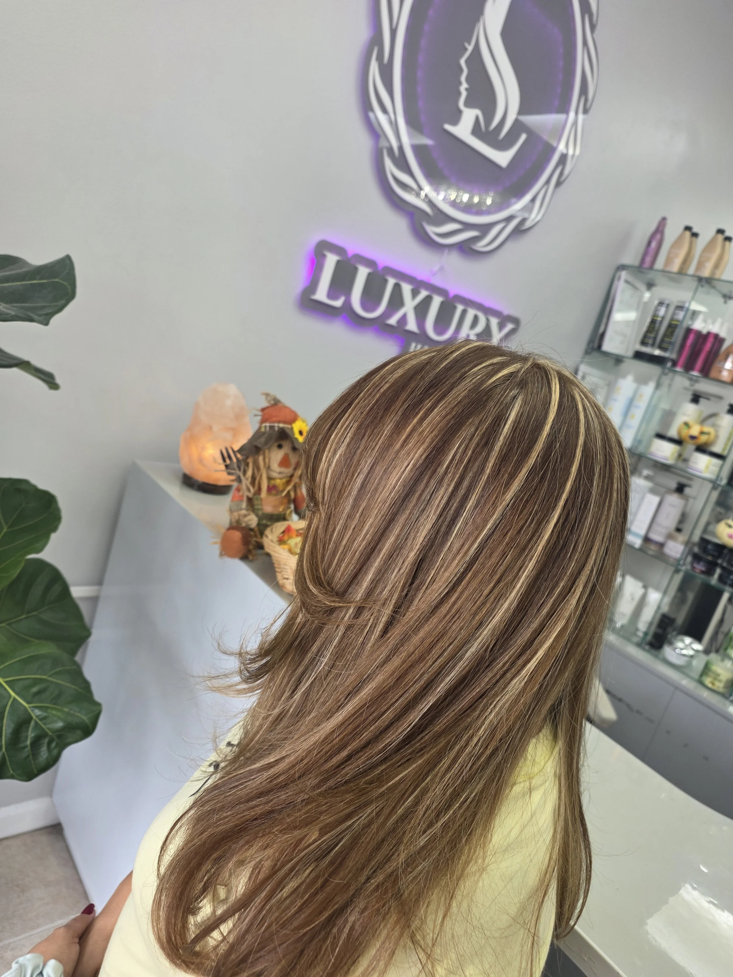 Close-up of a woman's long hair with light brown and blonde highlights, in a salon with white walls, a decorative mirror with purple lighting, shelves with hair products, and a salt lamp.