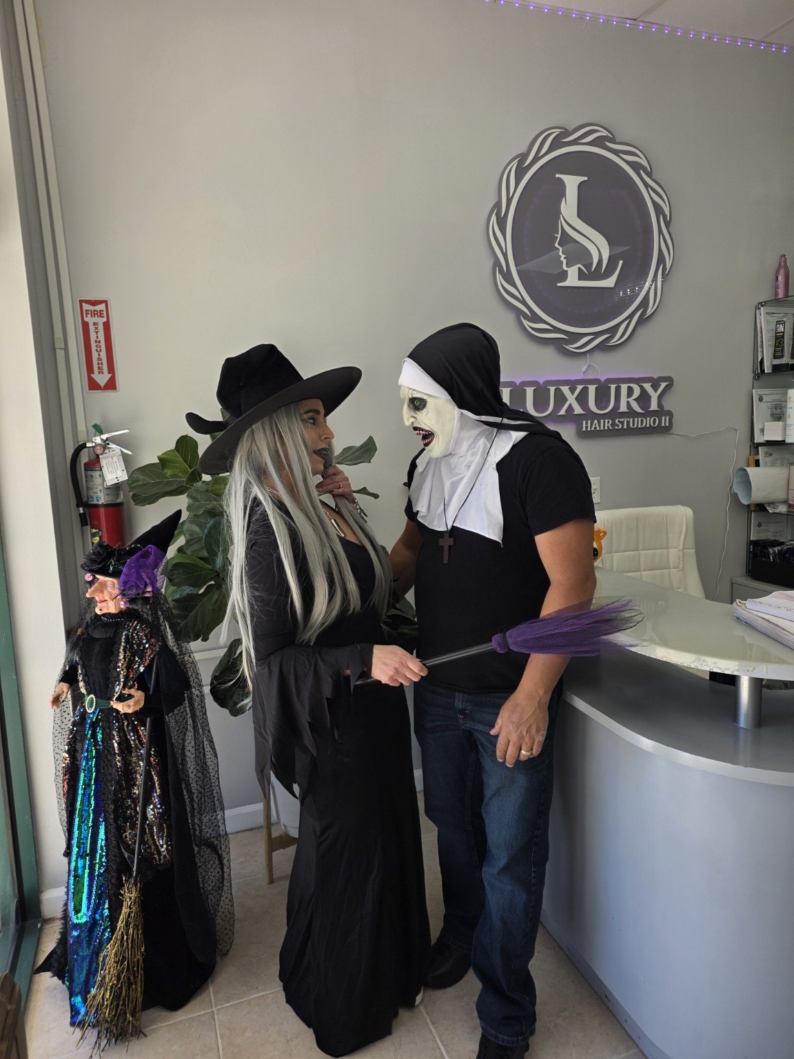 Two people dressed in Halloween costumes, one as a witch and the other as a nun, having a conversation inside a hair studio with a witch decoration on the side.