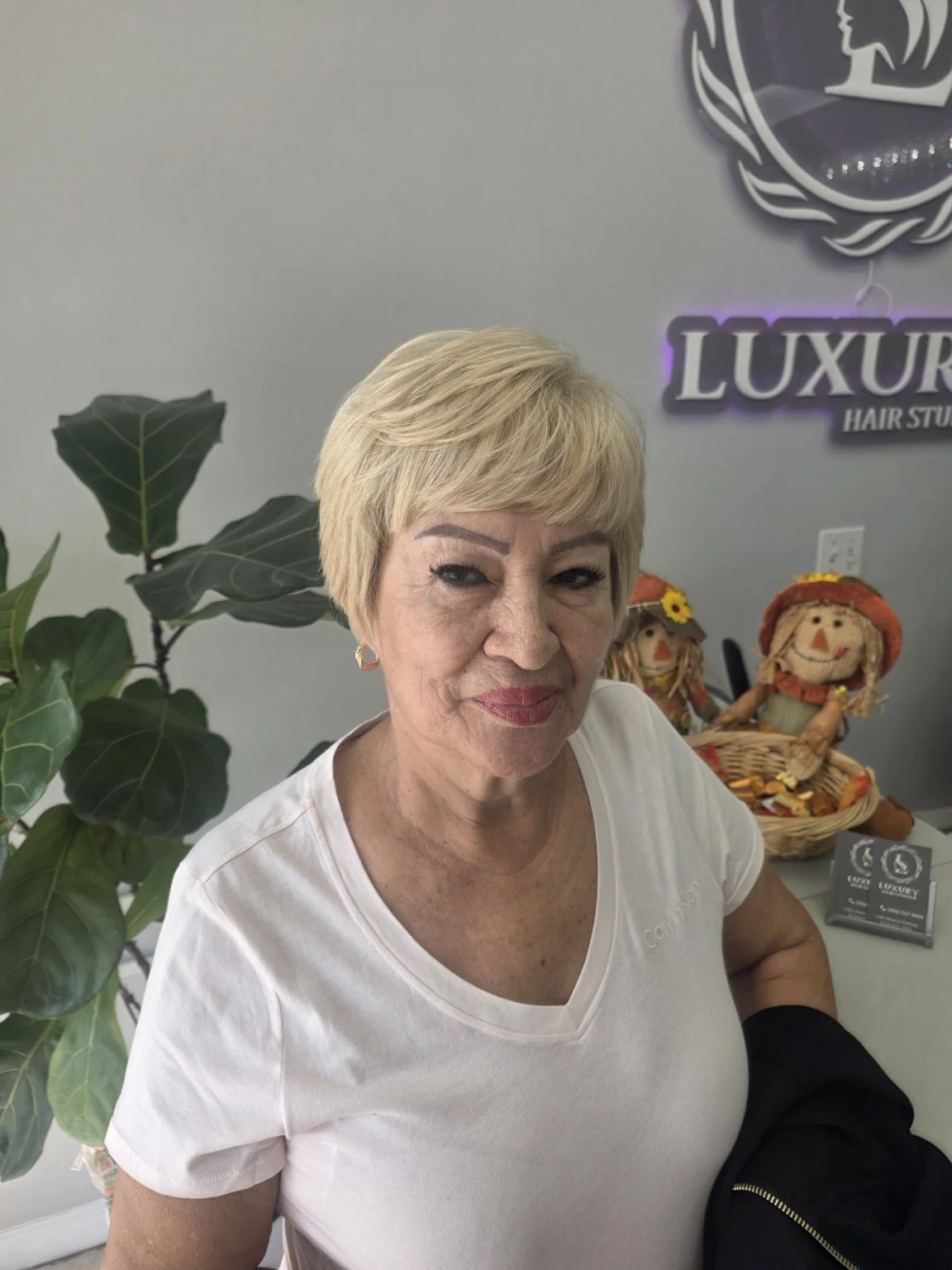 A woman with blonde hair, wearing a white shirt, standing inside a hair studio. Behind her are decorative dolls and a sign that says 'LUXURY Hair Studio'. There is a large green plant to her left.