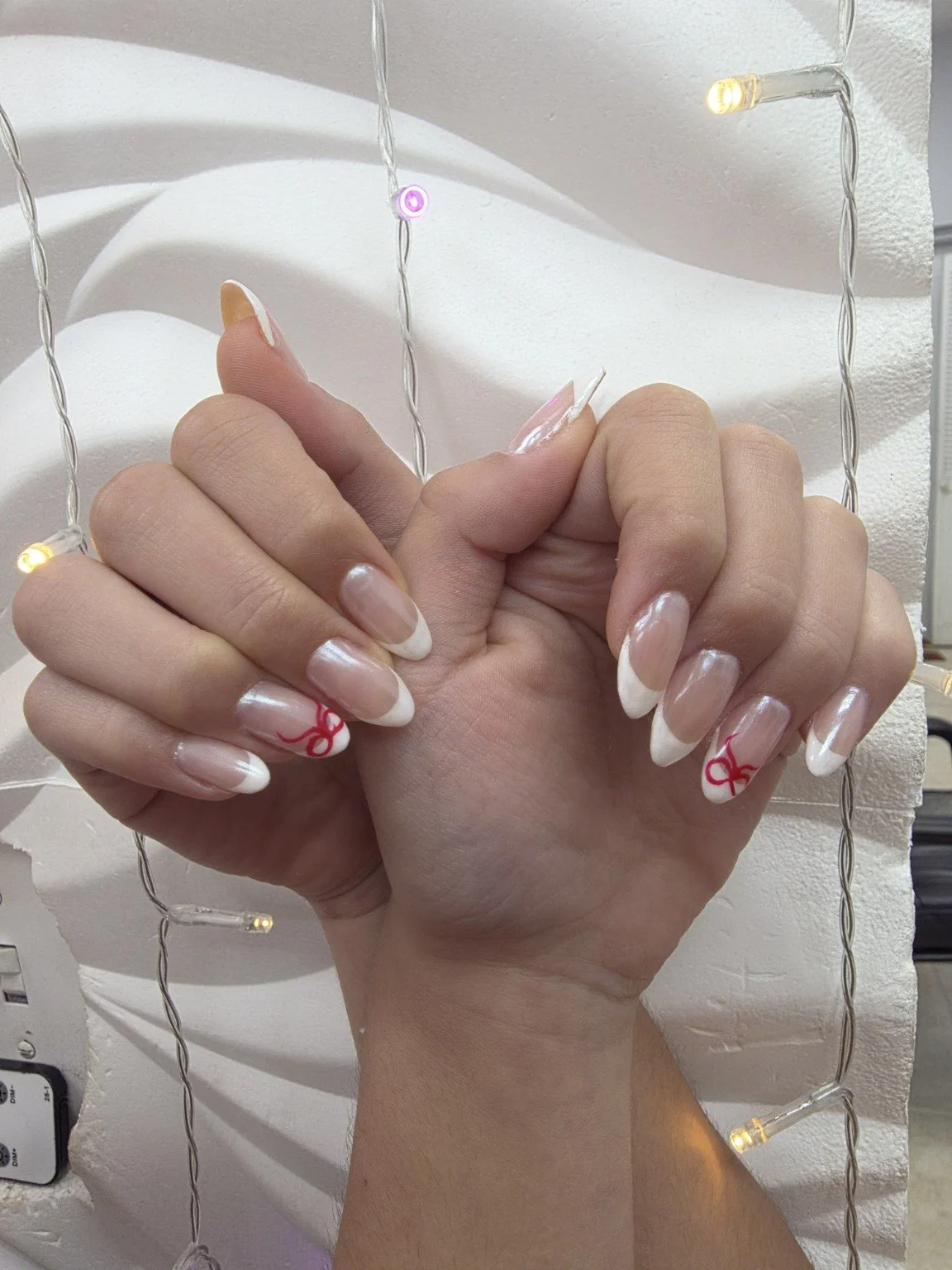 Close-up of a person's hand with acrylic nails painted with a natural base and white tips, featuring red bow designs on the ring fingers, against a white textured background with string lights.