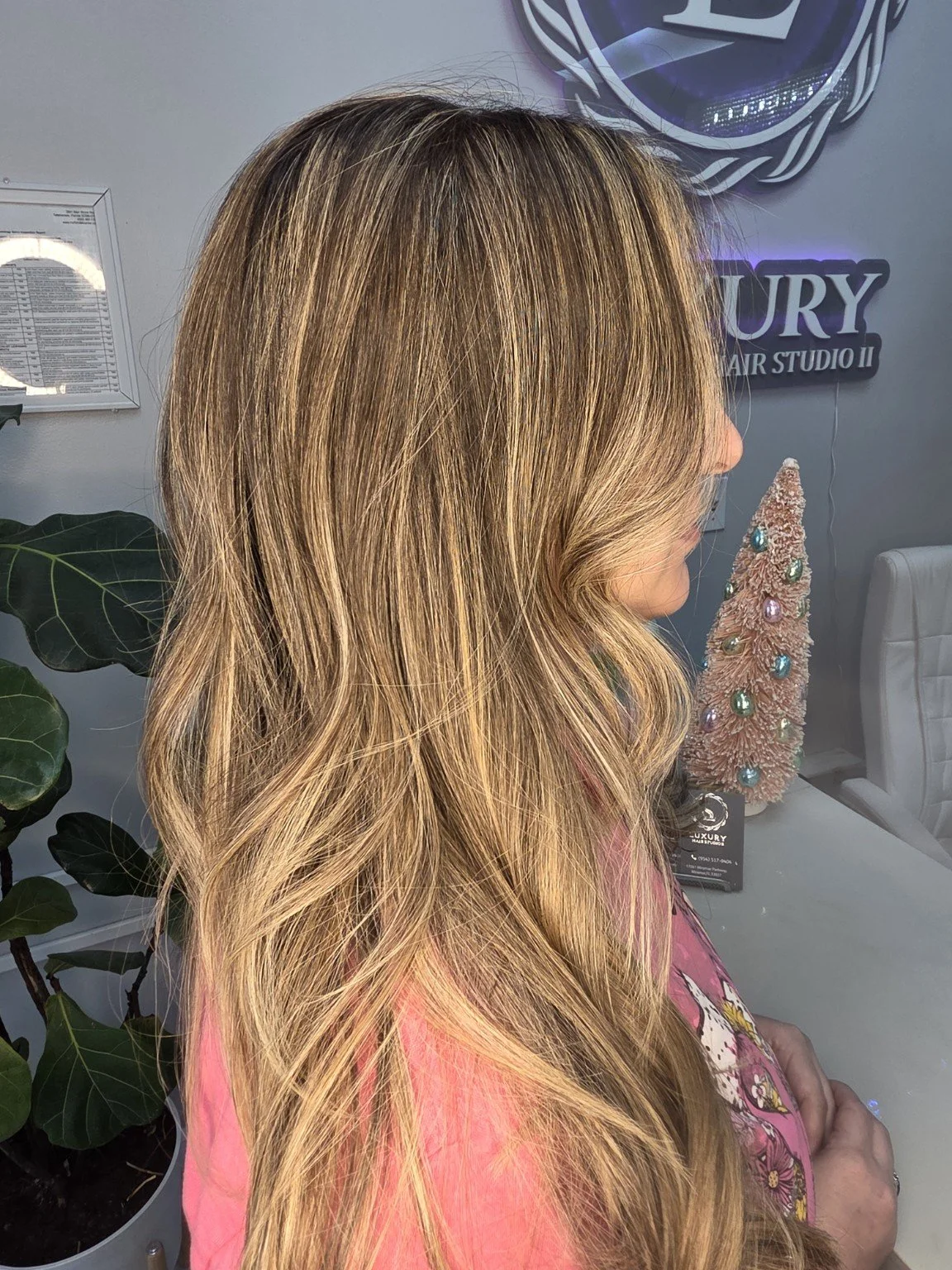 Side view of a woman with long, wavy, blonde hair standing indoors near a decorative Christmas tree and a wall with a sign that reads 'LUXURY HAIR STUDIO II'.