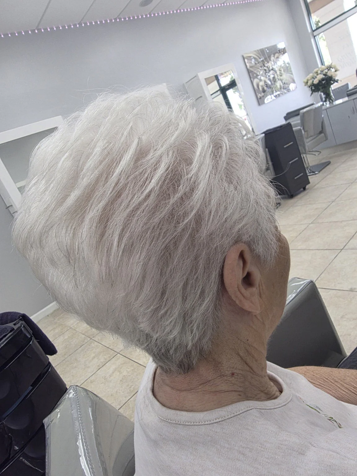Close-up side view of an elderly woman with short, white hair sitting in a salon chair inside a hair salon.