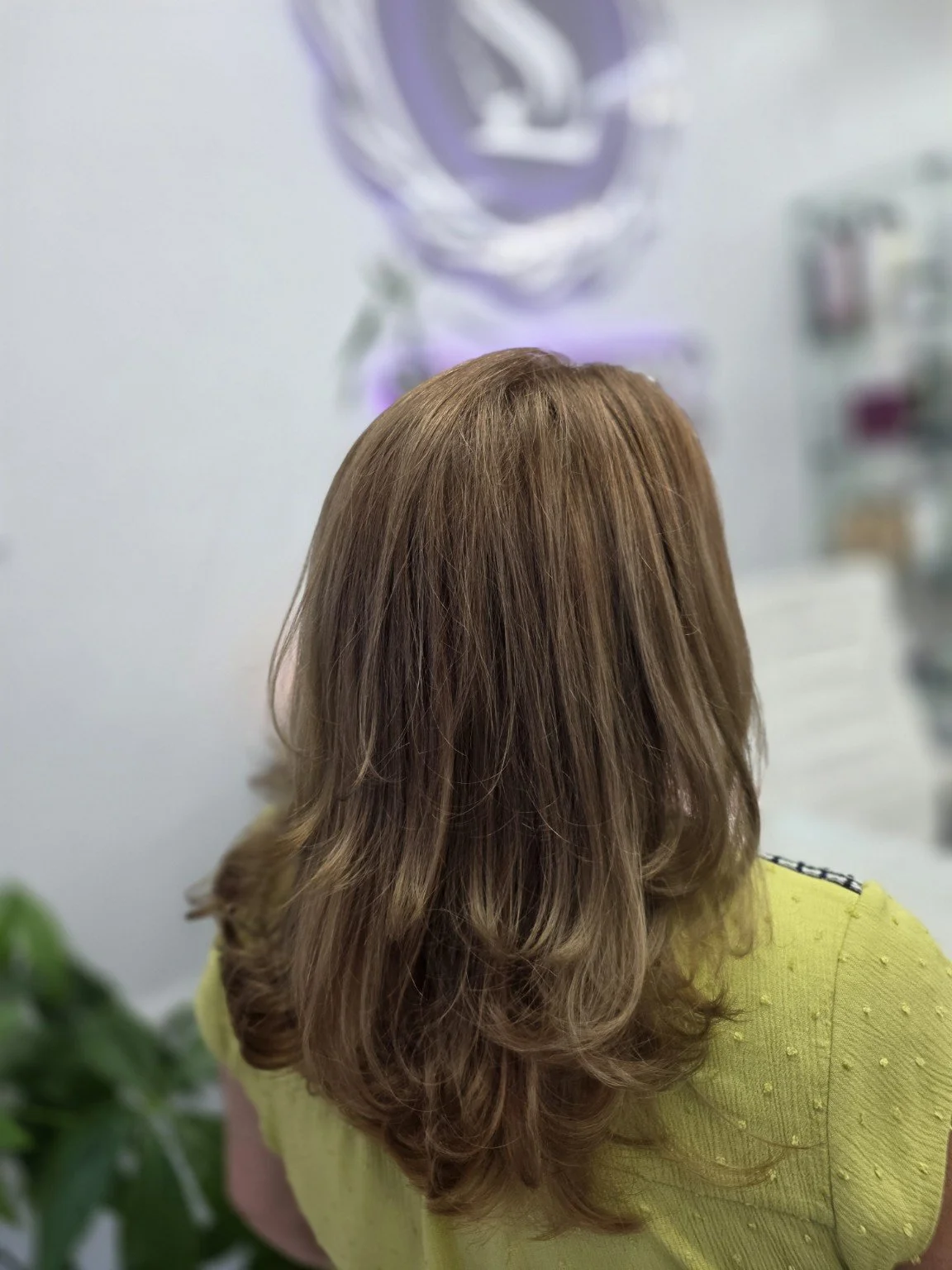 Back view of a woman with shoulder-length, wavy light brown hair in a salon or store with a blurred background.