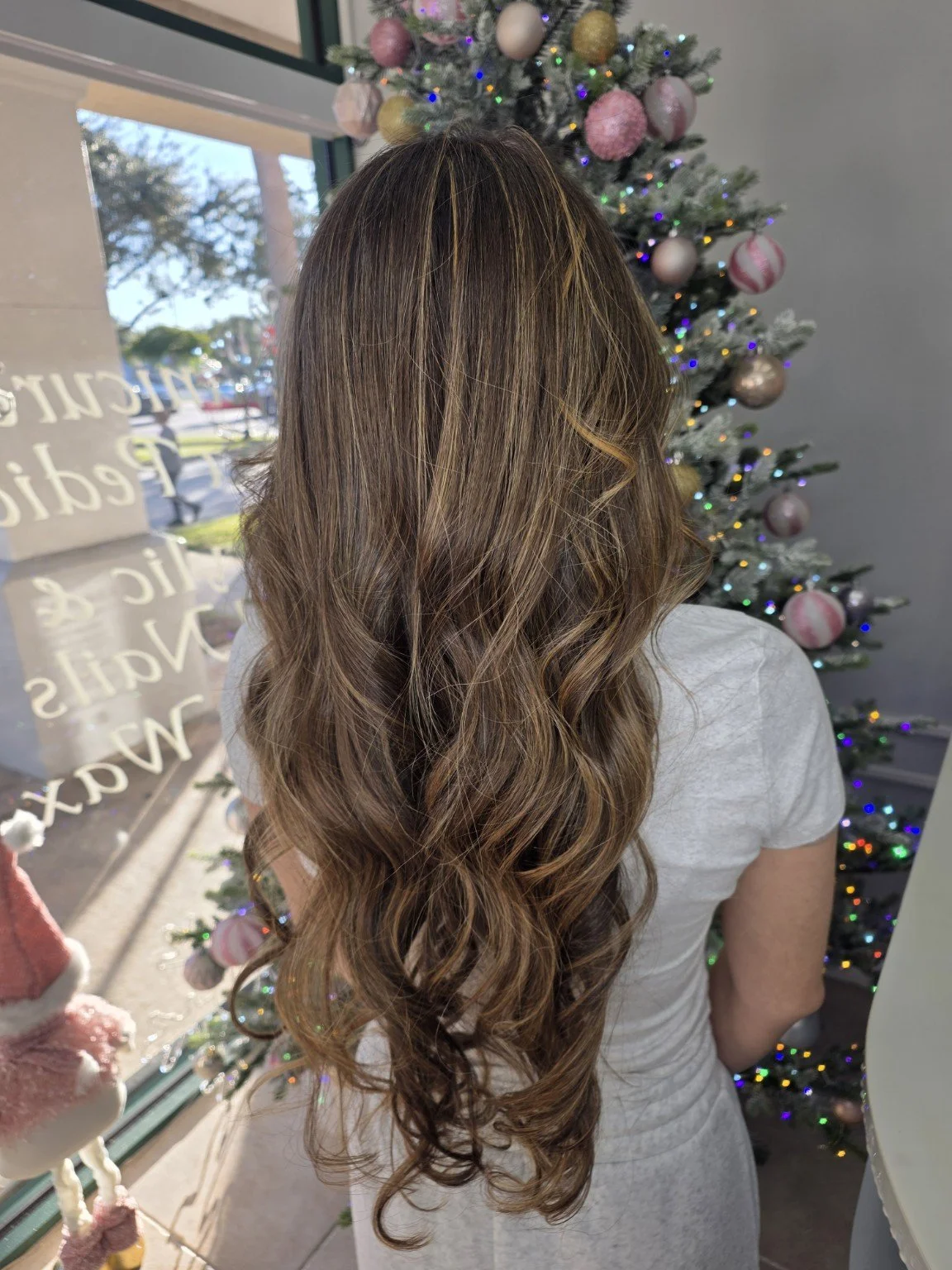 Back view of a woman with long, wavy, light brown hair near a decorated Christmas tree, with a window in the background.