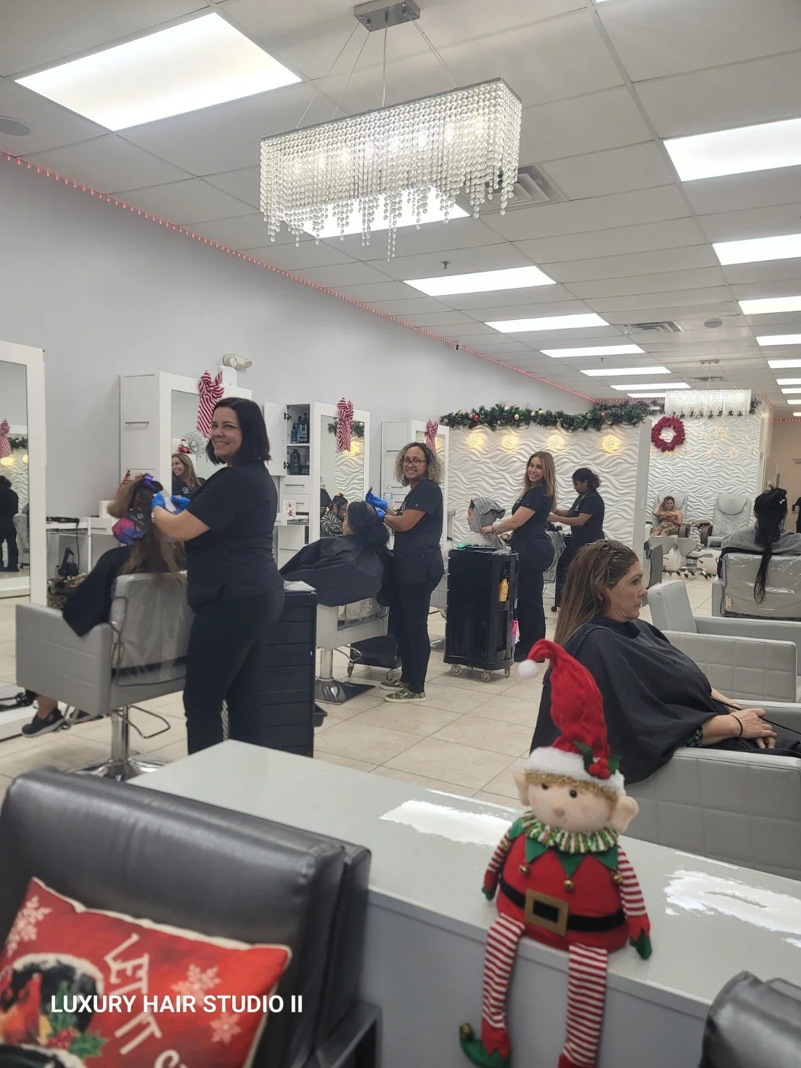 Interior of a hair salon decorated for Christmas with barbers and clients, some with Santa hats, and festive decorations including wreaths and garlands.