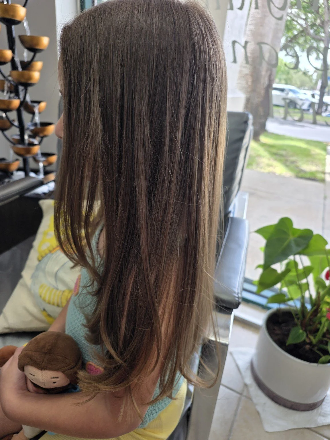 A woman with long, straight brown hair is seated inside a salon or similar setting near a window with green trees outside. She holds a plush toy in her hand. The background features a potted plant and salon equipment.