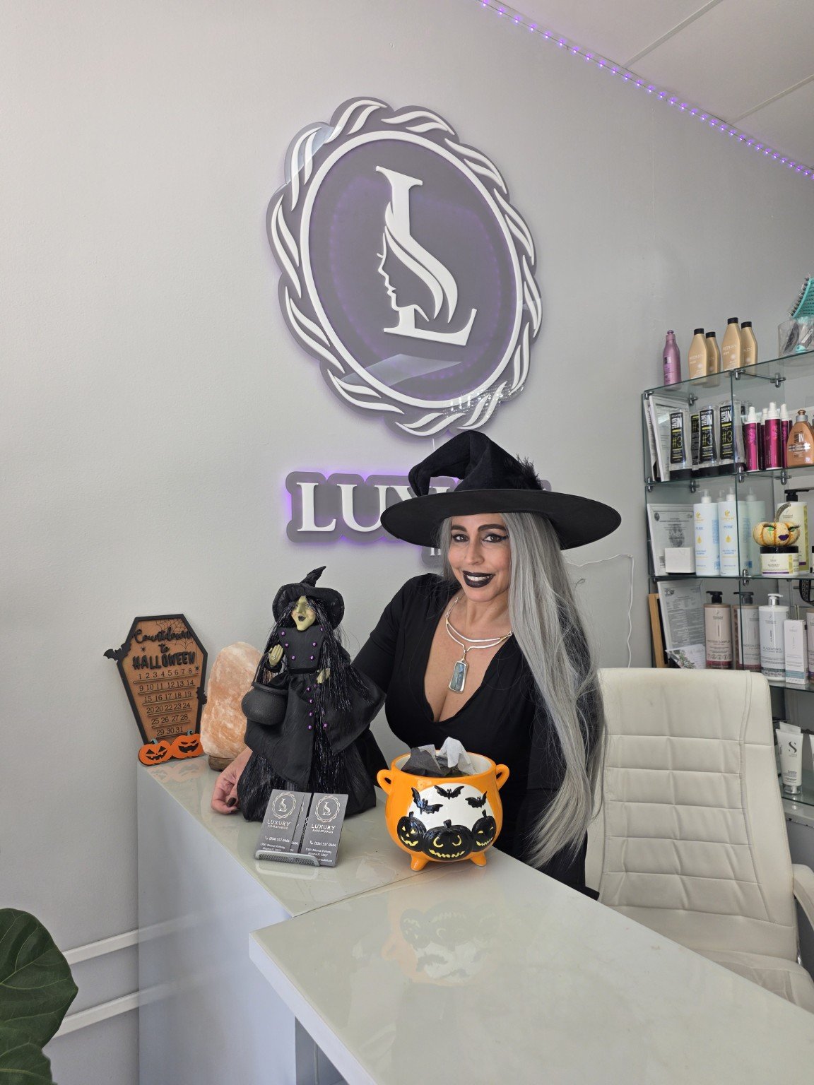 A woman dressed as a witch with gray hair and black makeup, wearing a witch hat, sitting behind a counter with Halloween decorations, including a witch doll, a pumpkin-shaped container, and a salt lamp. The background features a white wall with a log