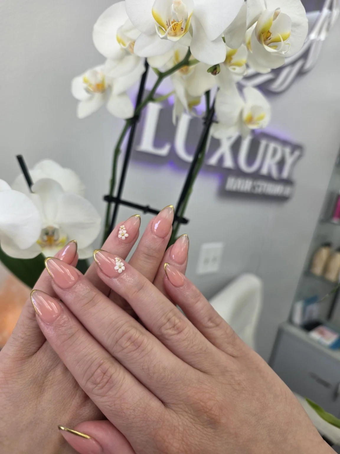 Close-up of hands with manicured nails and small white flower nail art, behind a display of white orchids and a sign that reads 'LUXURY'.
