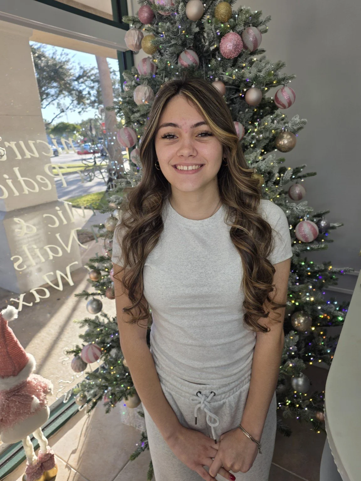 A young woman with long, wavy brown hair standing in front of a decorated Christmas tree, smiling at the camera inside a building.