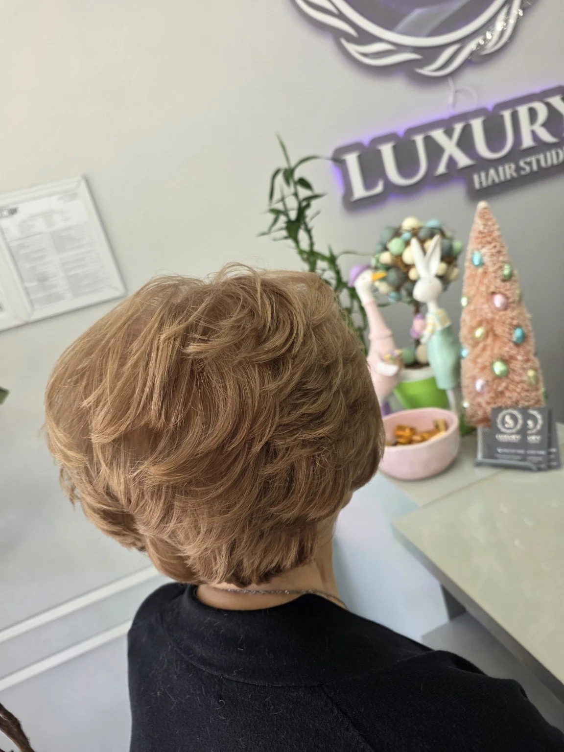 Side view of a woman with short, wavy, light brown hair, sitting at a table near a decorated display at a hair studio named 'Luxury Hair Studio.' The background features festive decorations, including a pink Christmas tree, a bunny figurine, and a si