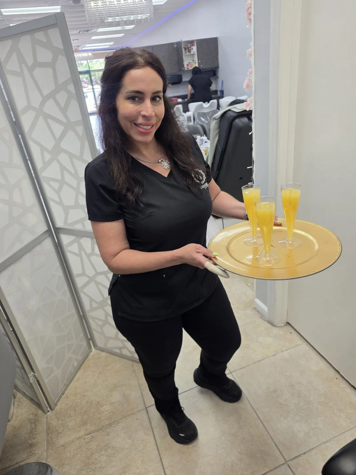 A woman in black clothing holding a silver tray with four tall glasses of yellow beverage inside a restaurant or cafe.