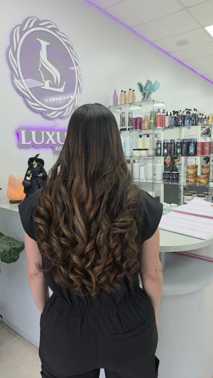 A woman with long, curly brown hair standing inside a hair salon with a sign that says 'LUXU' on the wall and shelves stocked with hair care products.