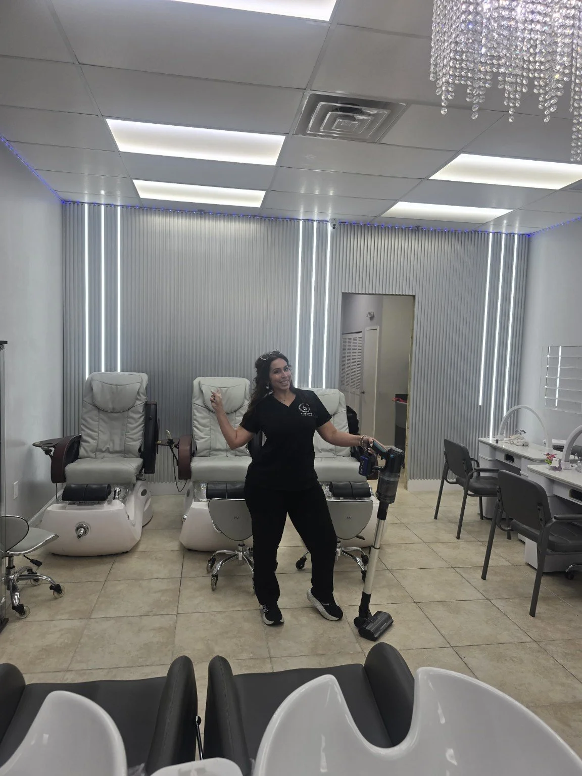 A woman in black scrubs smiling and holding a vacuum, standing in a medical or spa treatment room with three massage chairs, a desk with chairs, and decorative lighting on the back wall.
