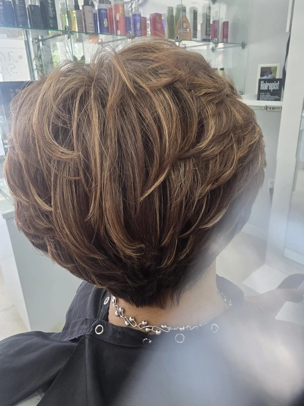 Back view of a person with short, layered, caramel and light brown highlighted hairstyle, sitting in a salon with hair products displayed in the background.