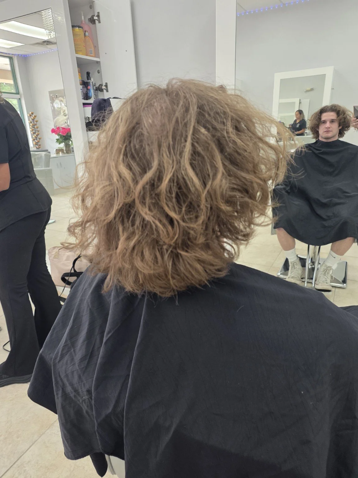 A person with curly, shoulder-length, light brown hair sitting in a salon chair, wearing a black smock, while a man with long, wavy, dark hair and casual attire sits in a salon chair in front of a mirror in the background.