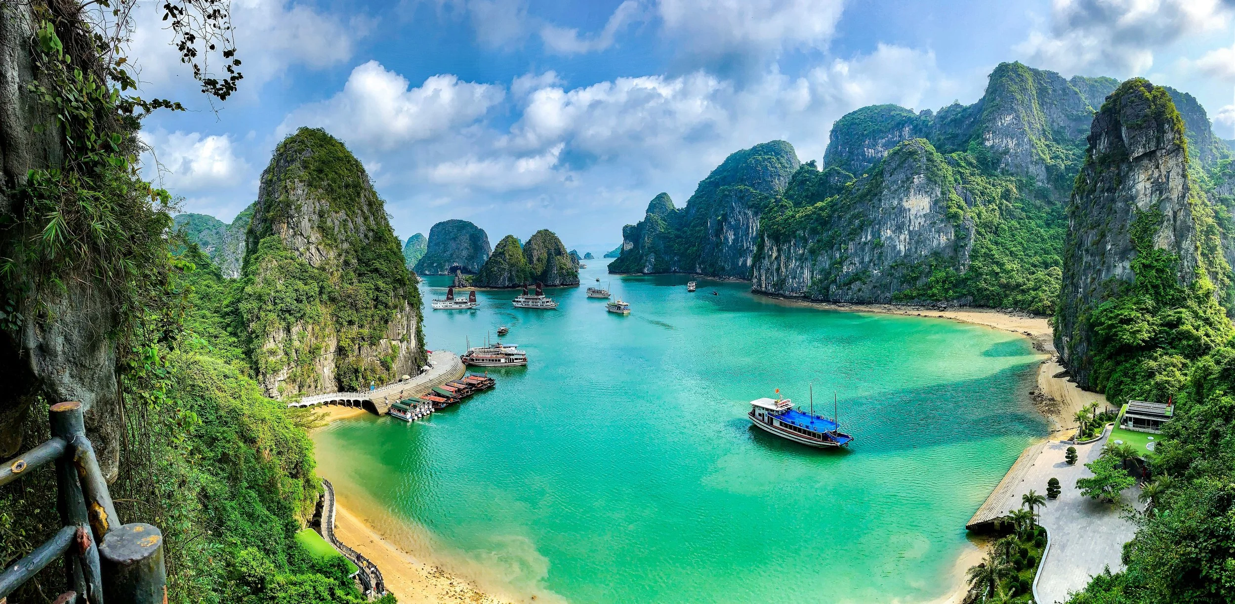 Scenic view of a bay with turquoise waters, surrounded by lush green limestone karsts and islands, with boats and ships in the water, and a small beach area on the right.