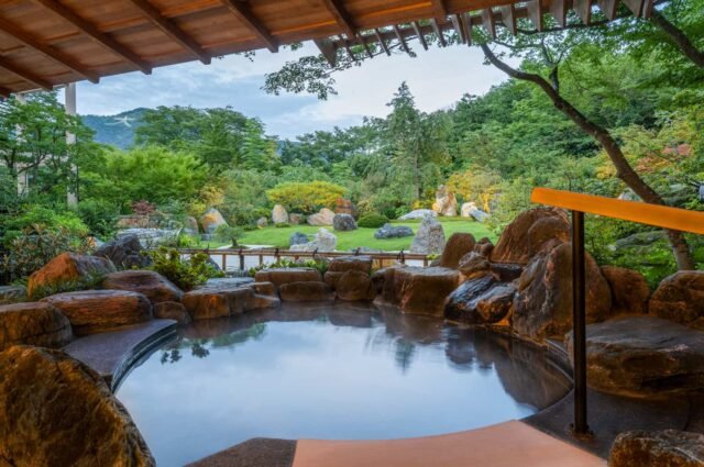Hot spring pool surrounded by rocks and lush green trees, viewed from a covered outdoor area.